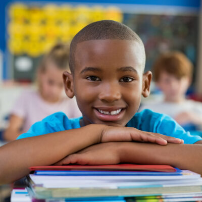 Portrait of african american schoolboy leaning on desk with classmates in background. Happy young kid sitting and leaning chin on stacked books in classroom. Portrait of elementary pupil looking at camera.